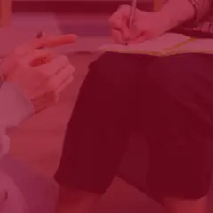 Dwie osoby siedzą naprzeciwko siebie, jedna pisze w notatniku, druga wskazuje palcem.