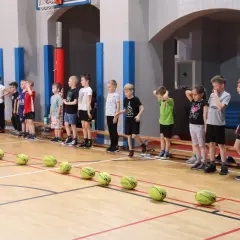 Dzieci w wieku 4-6 lat stoją w rzędzie na sali gimnastycznej, gotowe do gier i zabaw ruchowych. Na podłodze leżą piłki.
