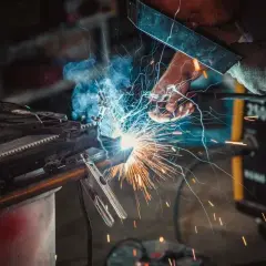 Spawacz w masce tworzy iskrzący łuk, łącząc metalowe elementy. W tle żółta spawarka, a wokół latają iskry.