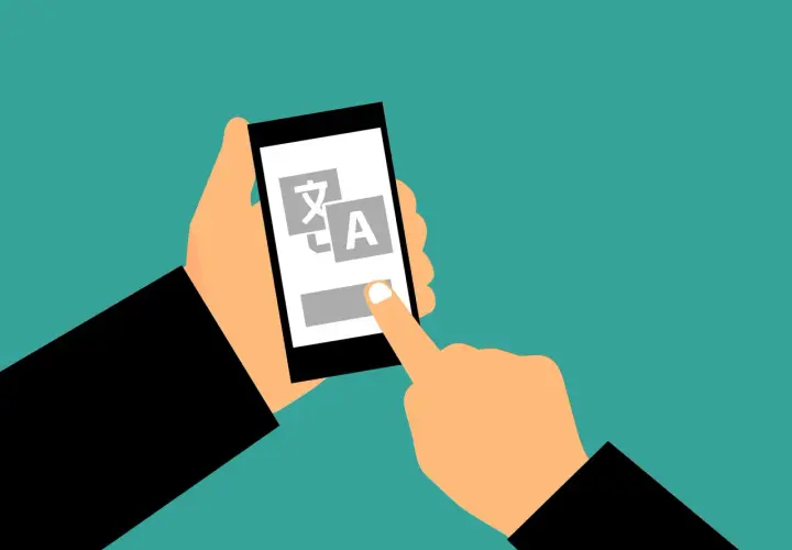 Dłoń trzyma smartfon z ikonami języków, a palec dotyka ekranu. Także czy tak że, tłumaczenie jest na wyciągnięcie ręki.