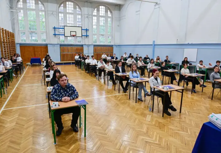 Uczniowie siedzą w ławkach w sali gimnastycznej, zdając egzamin. To typowy widok, gdy młodzi ludzie w wieku ok. 18 lat zdają maturę.