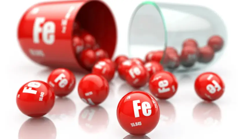 Czerwona kapsułka rozsypuje małe kuleczki z symbolem "Fe" i liczbami, symbolizujące żelazo.