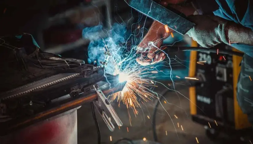Spawacz w masce tworzy iskrzący łuk, łącząc metalowe elementy. W tle żółta spawarka, a wokół latają iskry.