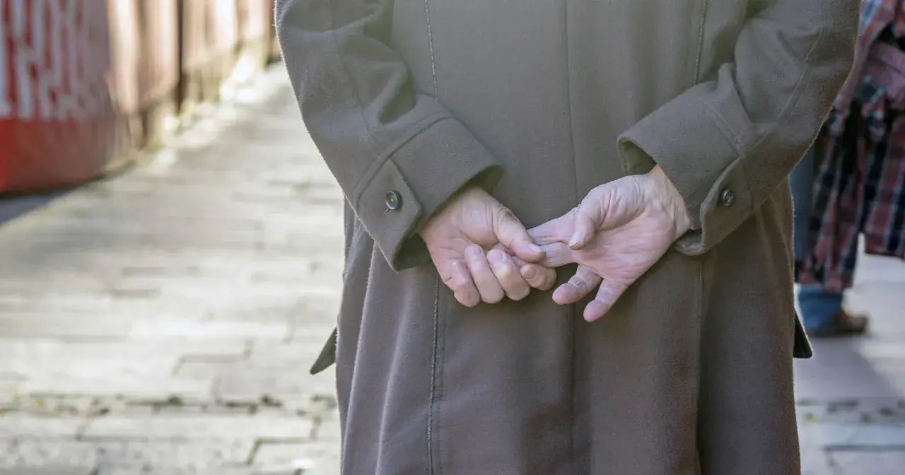 Osoba w brązowym płaszczu, z rękami splecionymi za plecami, stoi na chodniku.