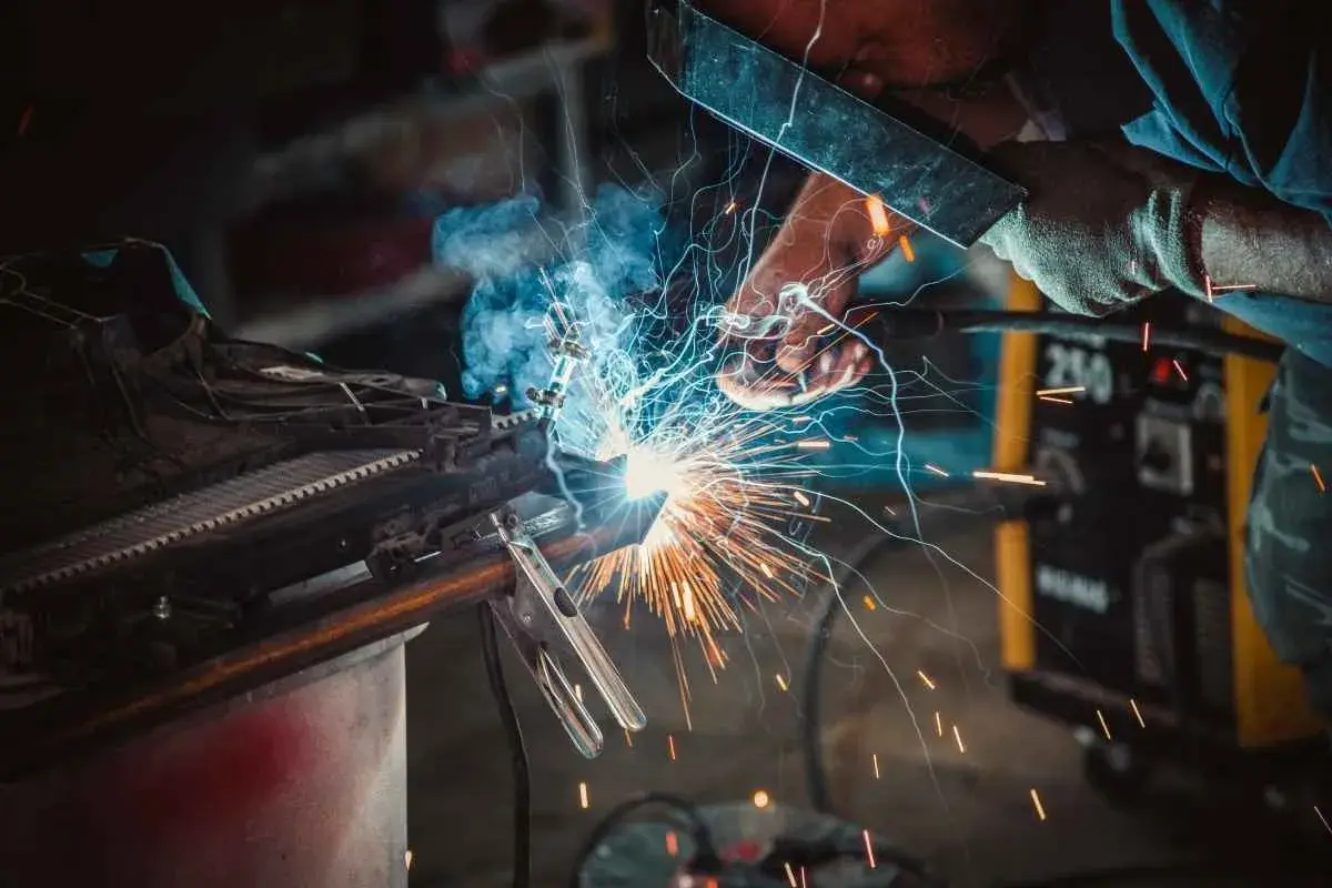 Spawacz w masce tworzy iskrzący łuk, łącząc metalowe elementy. W tle żółta spawarka, a wokół latają iskry.