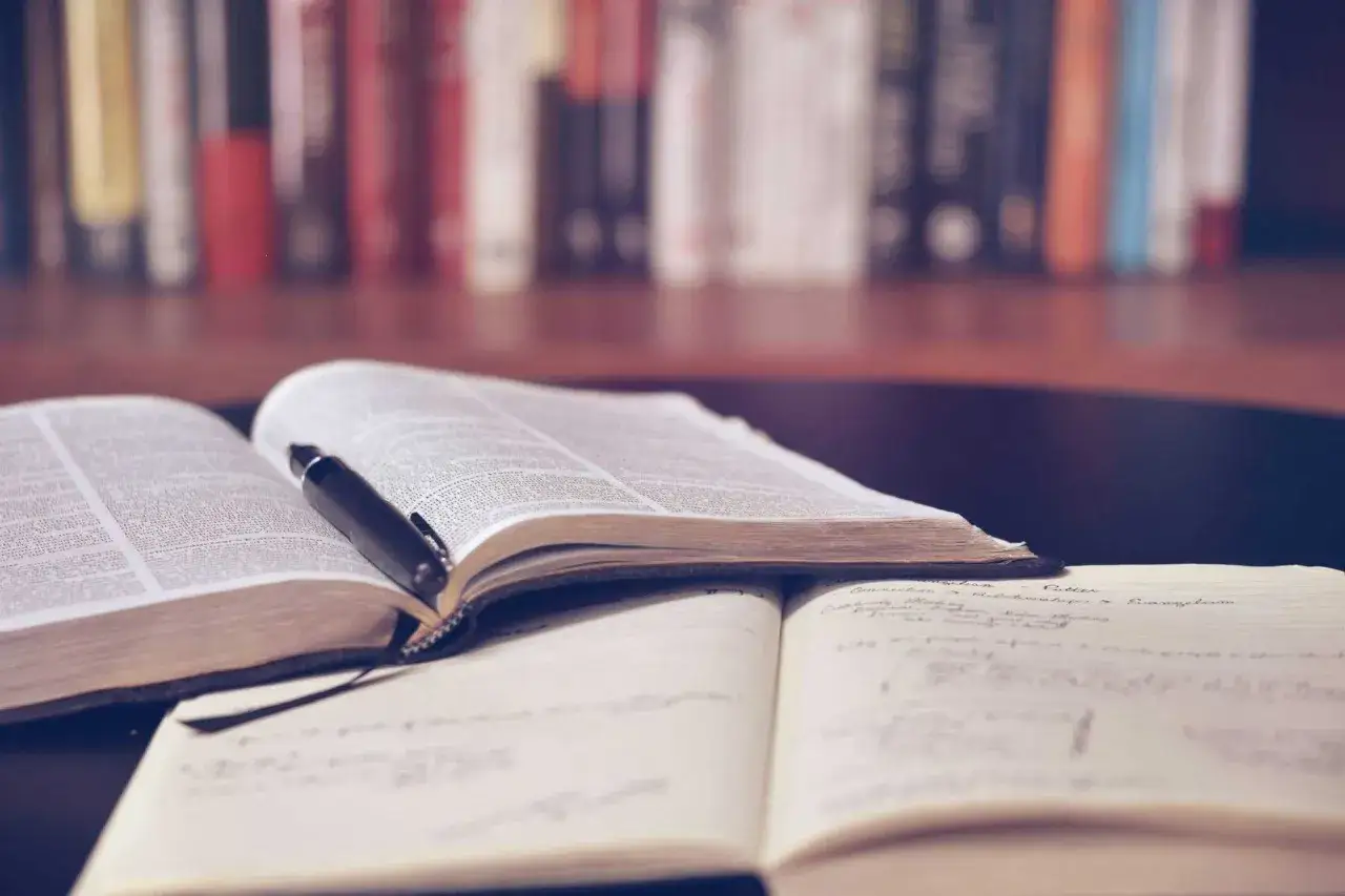 Otwarta księga z piórem, notatnik i półka z książkami w tle. Pedagogika rewalidacyjna zajmuje się wspieraniem rozwoju osób z trudnościami.