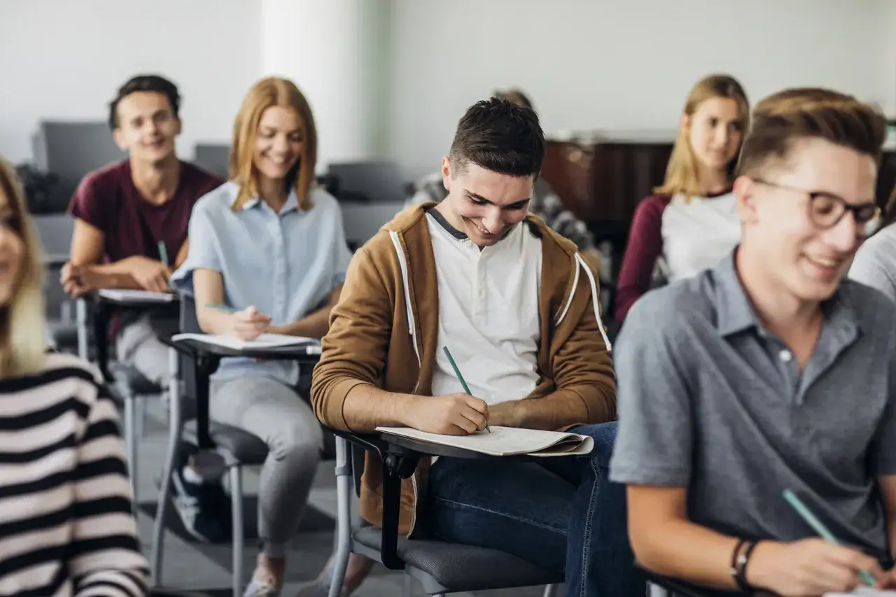 Uśmiechnięci studenci na lekcji. Czy to miłość od pierwszego wejrzenia, czyli crush? Chłopak w brązowej bluzie pisze coś w zeszycie.